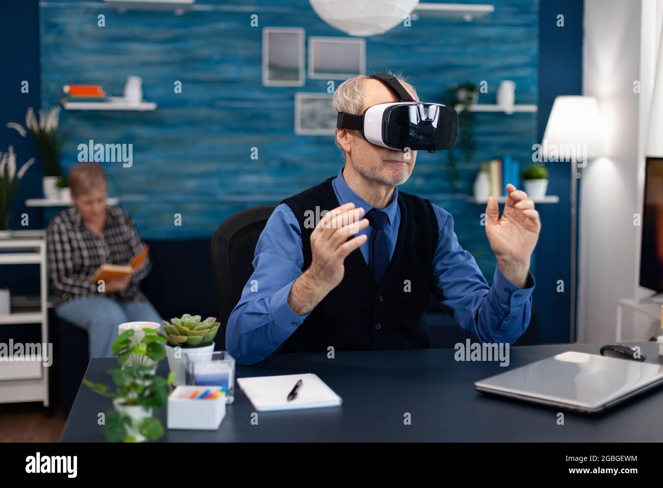 Heureux titulaire de l'offre de réalité virtuelle portant un micro-casque/une  oreillette. Homme âgé retraité dans la salle de séjour assis au bureau avec  des lunettes futuristes pour femme du cyberespace augmenté regardant