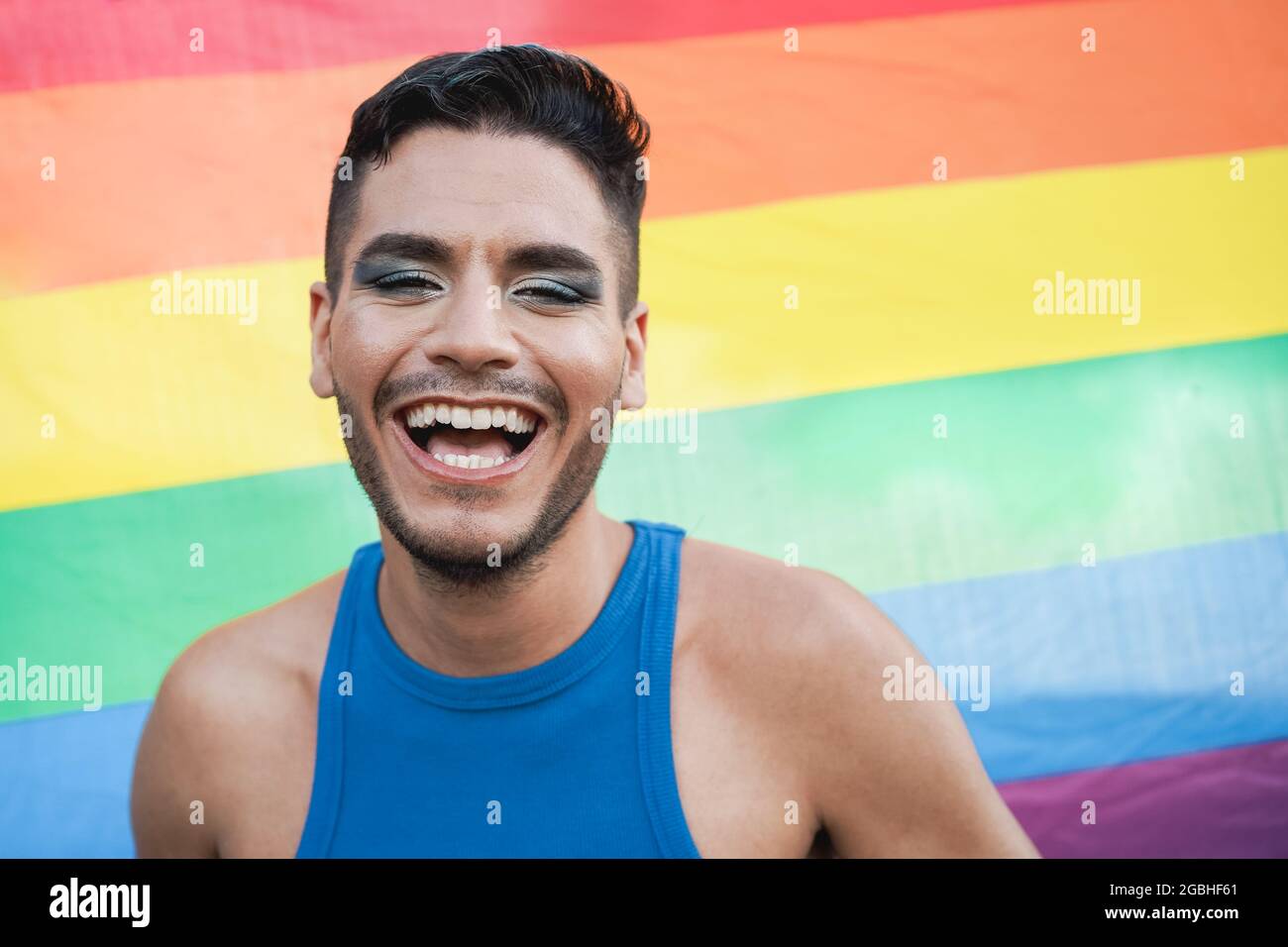 Jeune homme transgenre avec maquillage souriant sur la caméra avec le drapeau arc-en-ciel lgbt sur fond - Focus sur le visage Banque D'Images