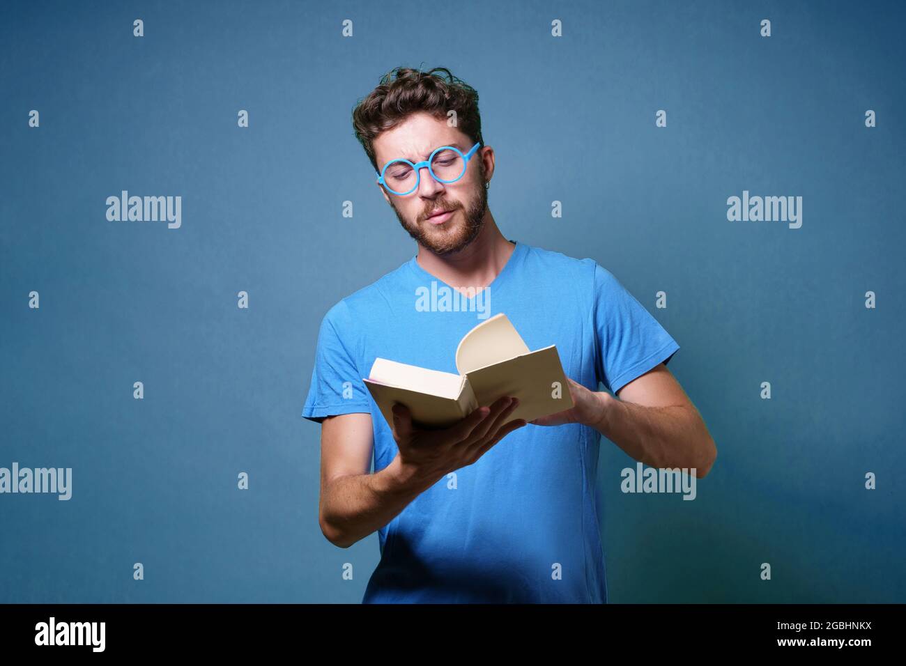 Portrait d'un jeune étudiant avec des lunettes lisant un livre. Portrait isolé sur fond bleu. Prise de vue en studio. Banque D'Images