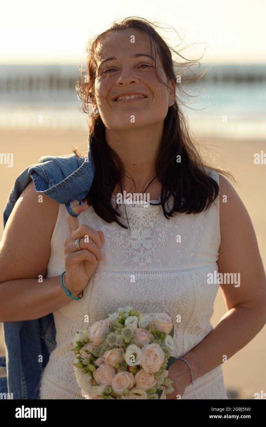 Heureuse femme moderne dans une élégante robe blanche avec une veste en denim accroché sur son épaule en souriant à l'appareil photo tout en portant une bouque de mariée Banque D'Images