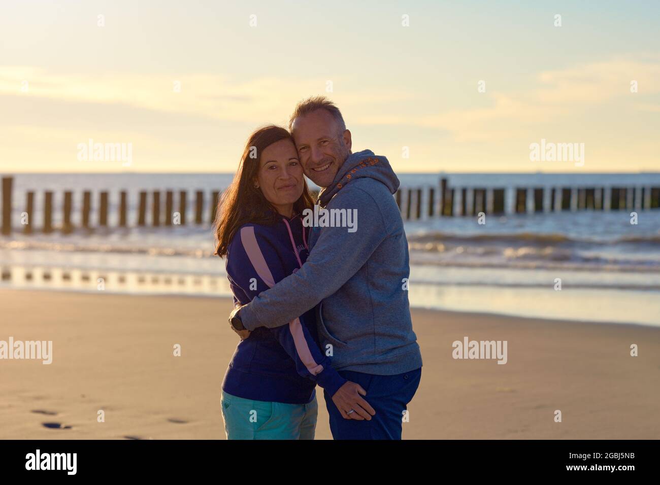 Joyeux couple romantique d'âge moyen qui profite d'un petit moment d'amour sur une plage tropicale déserte au coucher du soleil, debout dans un cadre intime et souriant à l'appareil photo wi Banque D'Images