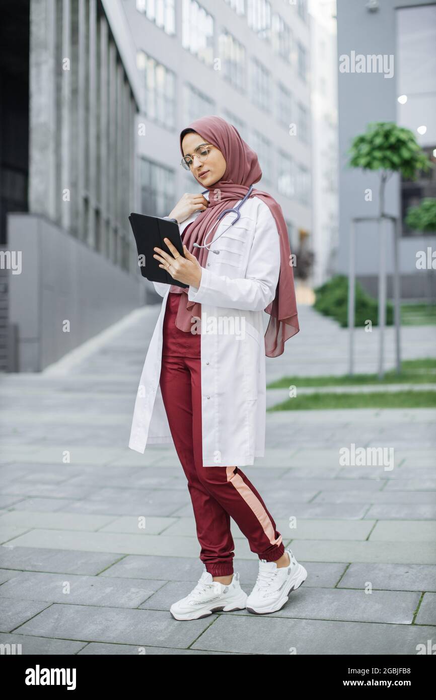 Femme musulmane médecin en hijab, portant des exfoliants et un manteau blanc, travaillant avec les dossiers des patients sur une tablette numérique en dehors de la clinique moderne Banque D'Images