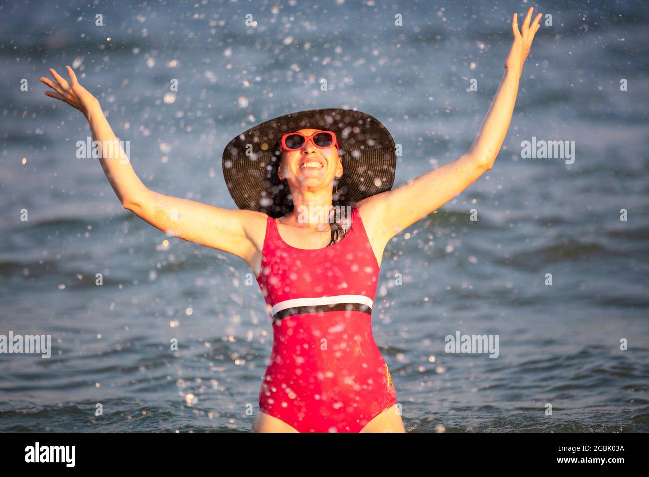 Femme heureuse à la mer dans le soleil. Femme dans un maillot de l'eau éclabousse sur fond de vagues. Vacances d'été sur la plage. Banque D'Images