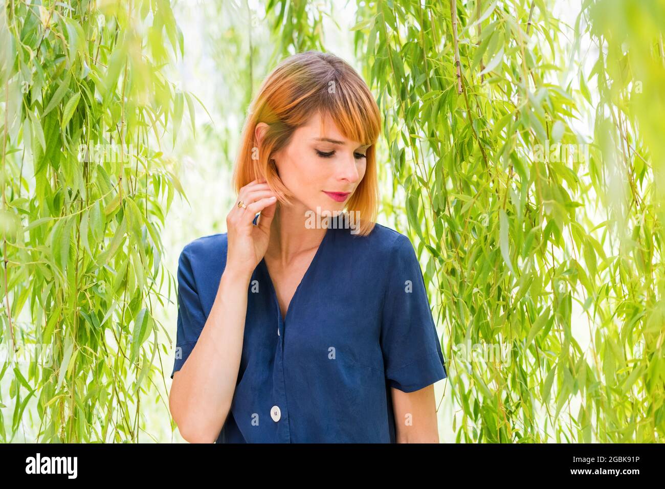 Portrait d'une belle femme sereine à tête rouge sous un saule pleurant regardant vers le bas avec une expression méditative tranquille et la main levée avec élégance à Banque D'Images