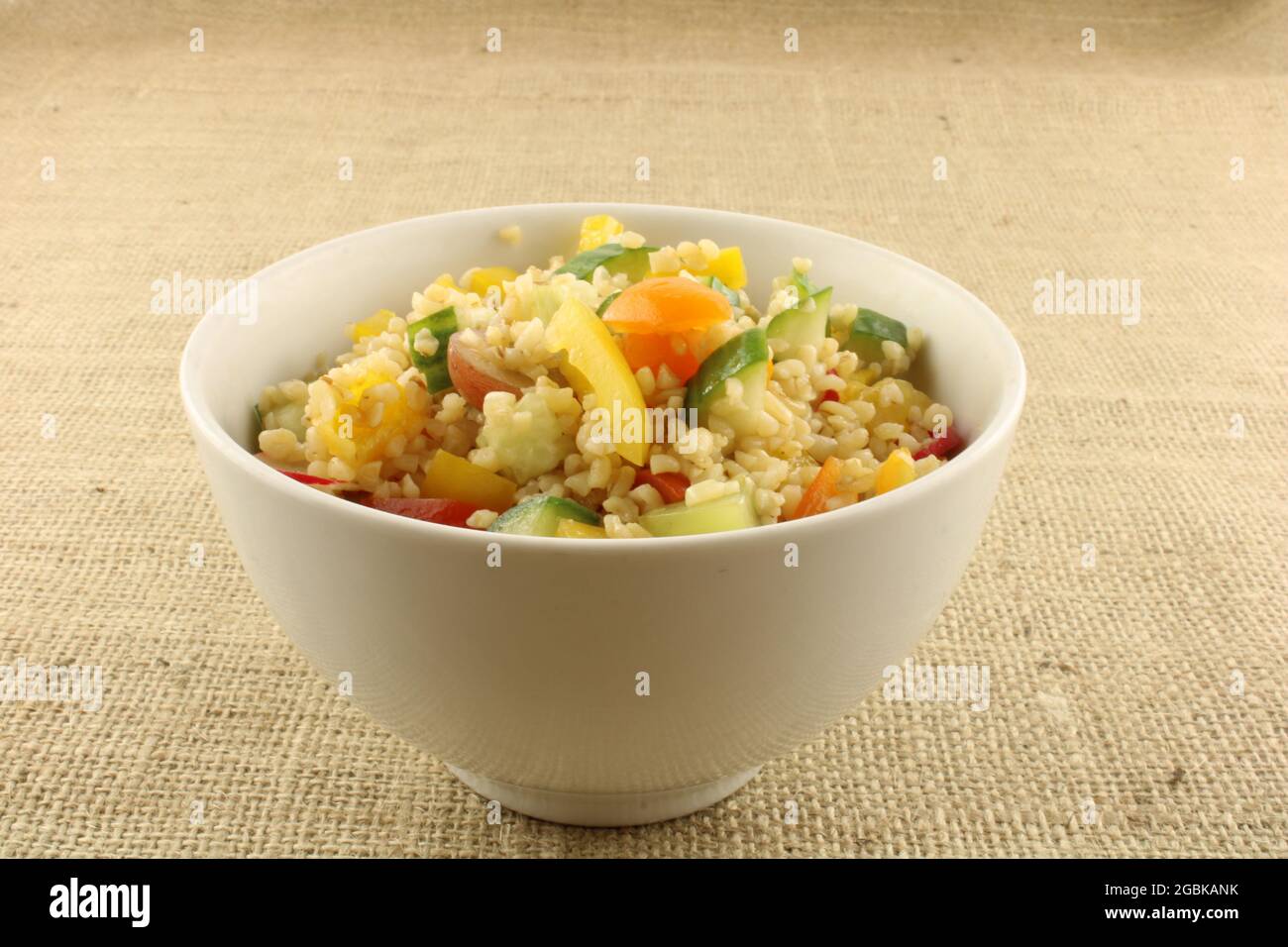Salade de Bulgur fraîchement préparée avec fruits et légumes colorés dans un bol blanc sur fond hessien brun Banque D'Images