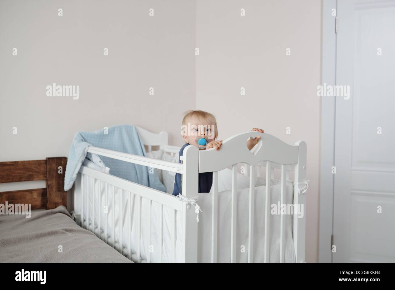 Portrait d'un bébé garçon actif qui suce un mannequin bleu et qui se tient dans un lit de bébé près du lit des parents Banque D'Images