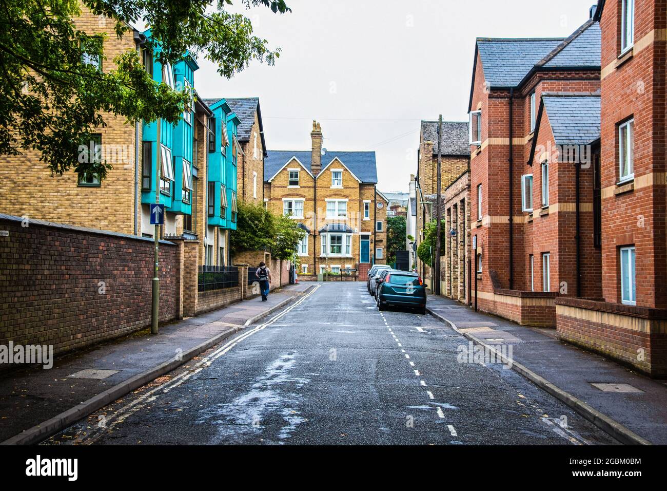 Oxford UK Street avec des voitures garées et de grands bâtiments de chaque côté et une chaussée mouillée et jeune mai avec sac à dos marche sur le trottoir - le panneau indique Ric Banque D'Images