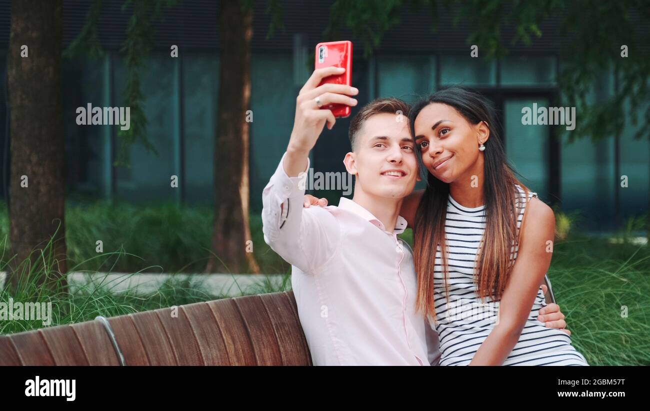 Jeune couple joyeux prenant selfie assis sur un banc à l'extérieur. Ils passent leur temps dans le grand parc. Banque D'Images