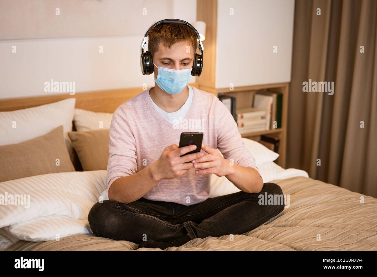 Un type de nationalité caucasienne en vêtements décontractés, portant un casque sur le lit dans un masque médical de protection avec un téléphone dans la main, regardant une vidéo en ligne sur un réseau Wi-Fi Banque D'Images
