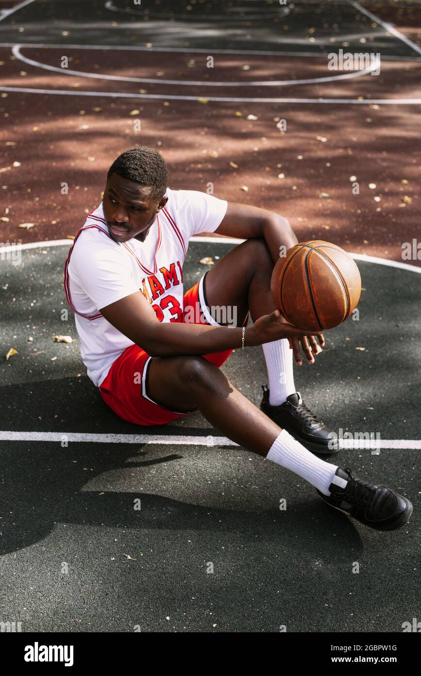 Portrait d'un jeune afro-américain tenant un ballon de basket et assis sur un terrain de basket-ball. Faites une pause pendant votre entraînement. Sport tendance Banque D'Images