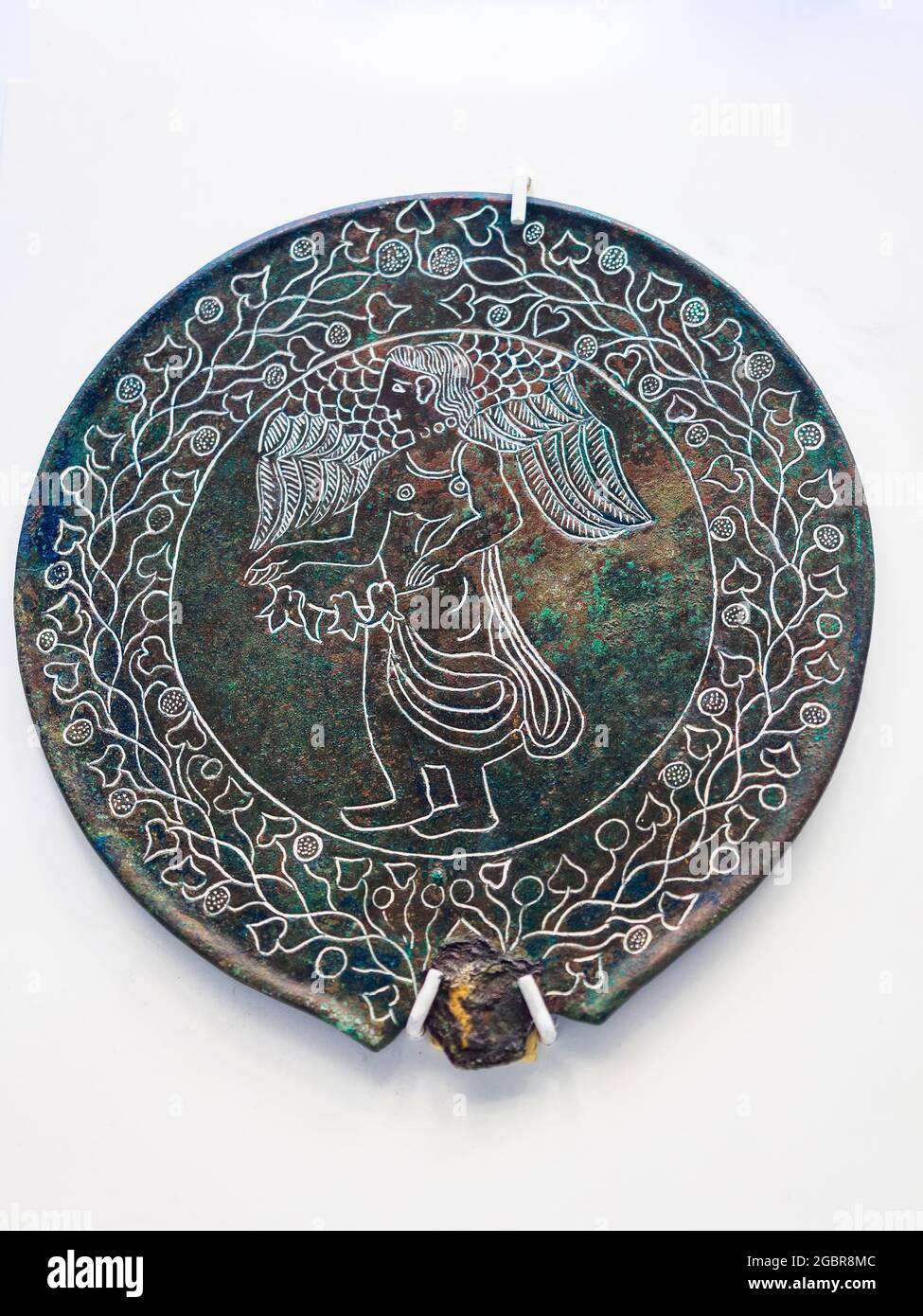 Miroir en forme de tenon circulaire étrusque représentant une figure femelle ailée face à gauche dans un cadre de guirlande de vignes et de corymbes portant un chiton qui descend vers les veaux et tient une guirlande de fleurs avec ses bras levés (5e cent BC) - Musée archéologique national de Tarquinia, Italie Banque D'Images