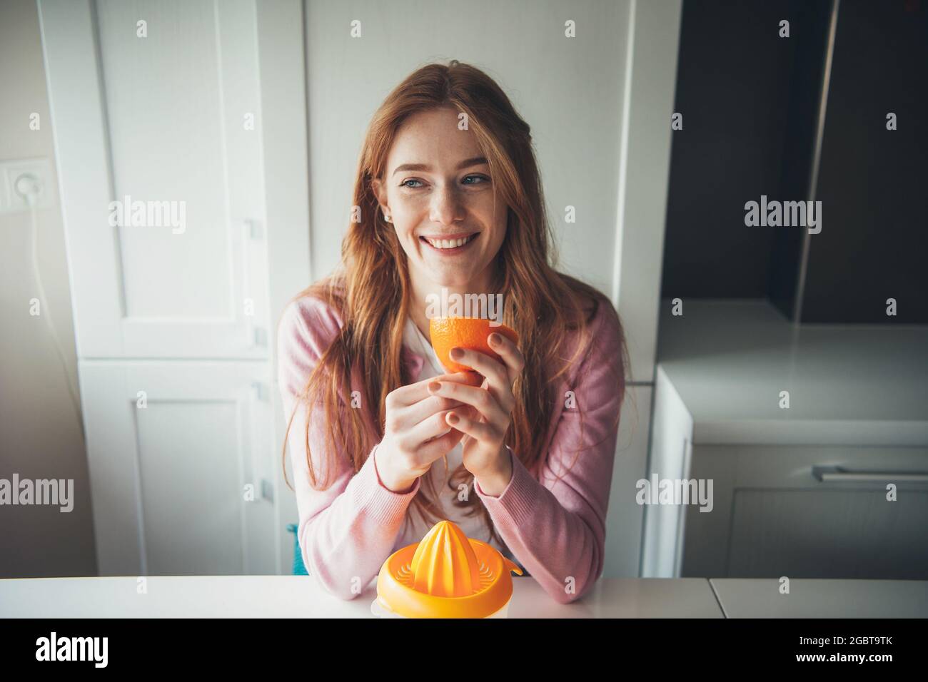 Ginger Woman avec des taches de rousseur sourit tout en tenant une tranche d'orange tout en comprimant le jus de fruit Banque D'Images