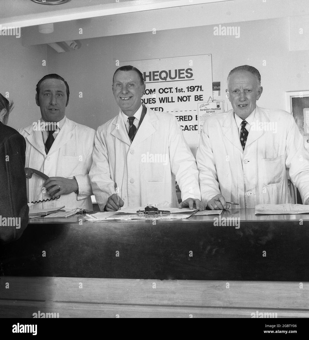1975, historique, à la réception d'un service de voiture, trois cadres supérieurs en manteaux blancs se tiennent derrière le comptoir, Angleterre, Royaume-Uni. Banque D'Images