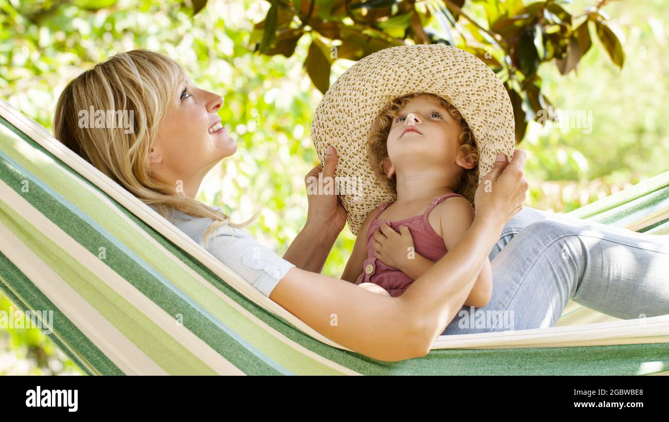 maman souriante et petite fille enfant yeux bleus avec des cheveux blonds bouclés, ensemble allongé sur le hamac dans le jardin vert de la maison, joue avec elle par Banque D'Images