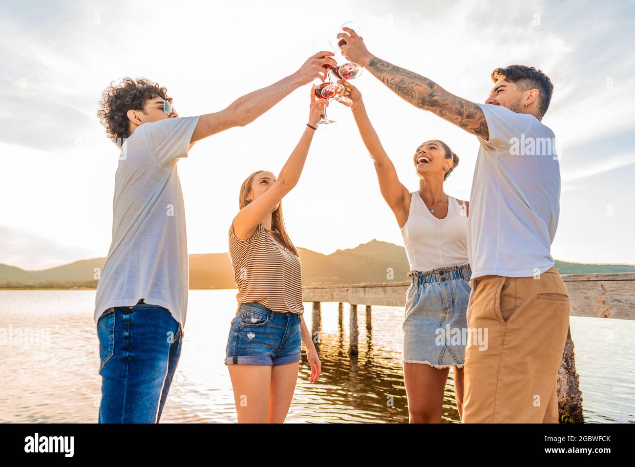 Groupe d'amis heureux multiraciaux en train de griller sur la plage avec du vin rouge au coucher du soleil verres à clinking devant la lumière du soleil en contre-jour. Les jeunes ont Banque D'Images