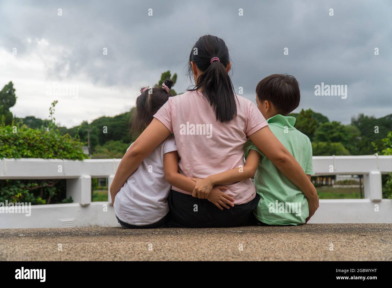 Vue arrière d'une mère asiatique aimante embrasser ses enfants assis sur le pont, la mère noire bienveillante embrasser l'enfant, se détendre en regardant le nuage noir, parent confort enfant Banque D'Images