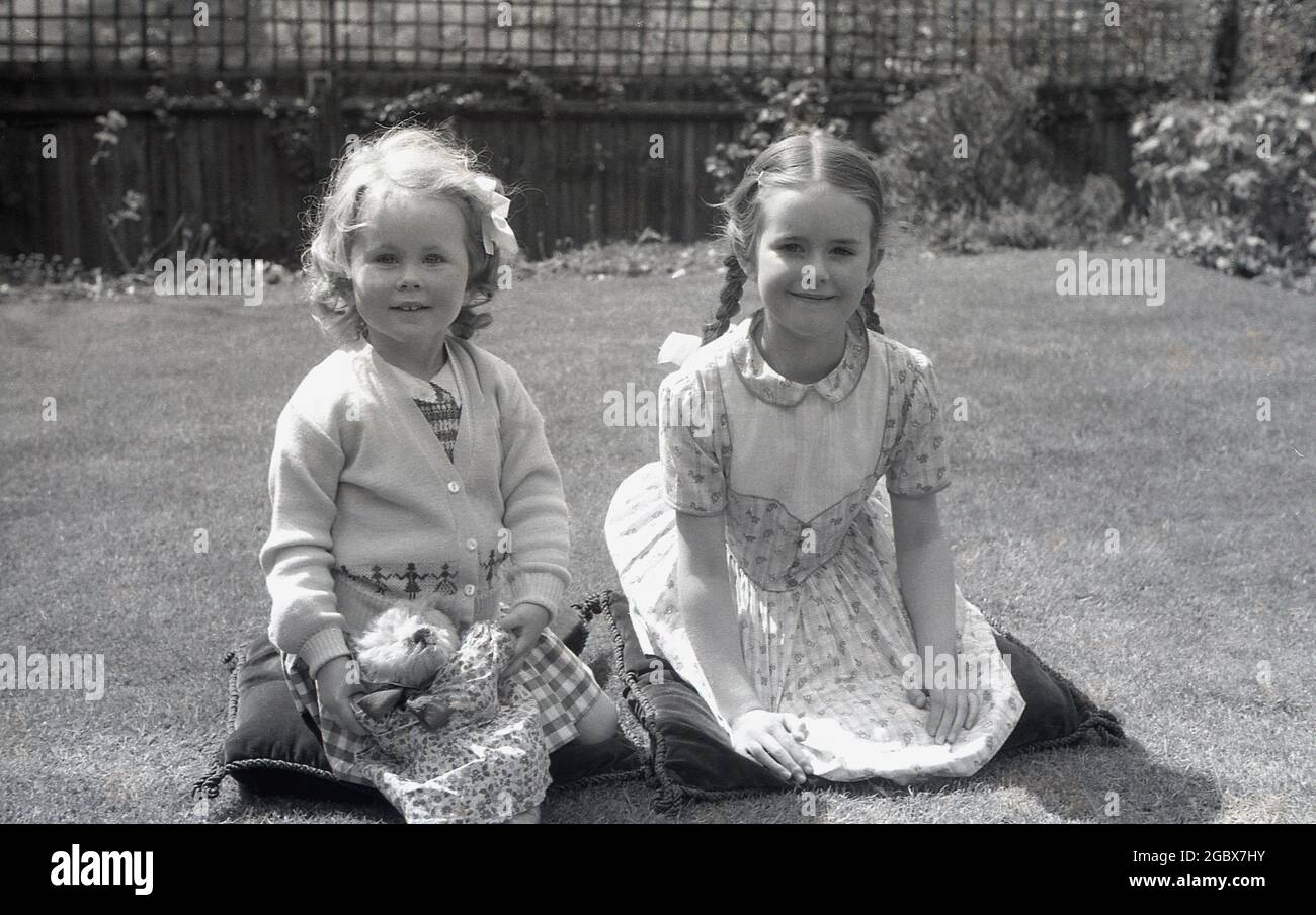dans les années 1960, deux jeunes filles historiques se tenaient ensemble dans un jardin sur des coussins pour leur photo, une tenant son jouet câleux, une petite teddy bear.in un papier d'emballage en coton. Banque D'Images