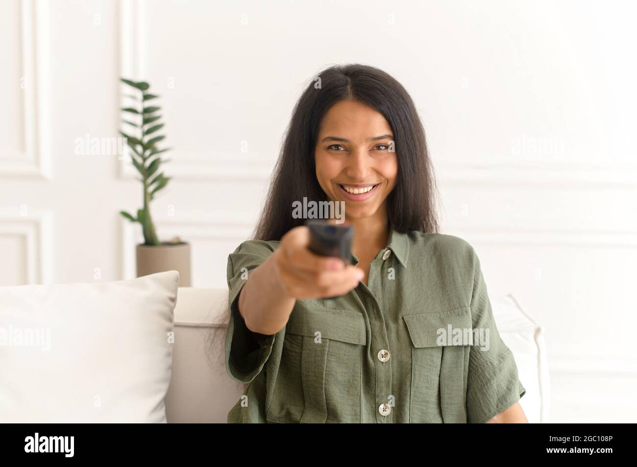 Belle jeune femme indienne regardant des programmes de télévision, assis sur un canapé confortable avec télécommande, changer de chaîne, regarde la caméra, multiraciale fille passe des loisirs à la maison Banque D'Images