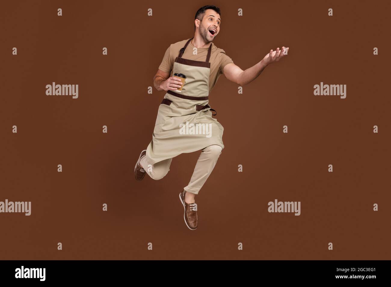 Homme pleine longueur de taille de corps portant un tablier sautant vers le haut en gardant tasse de café isolé couleur marron fond Banque D'Images