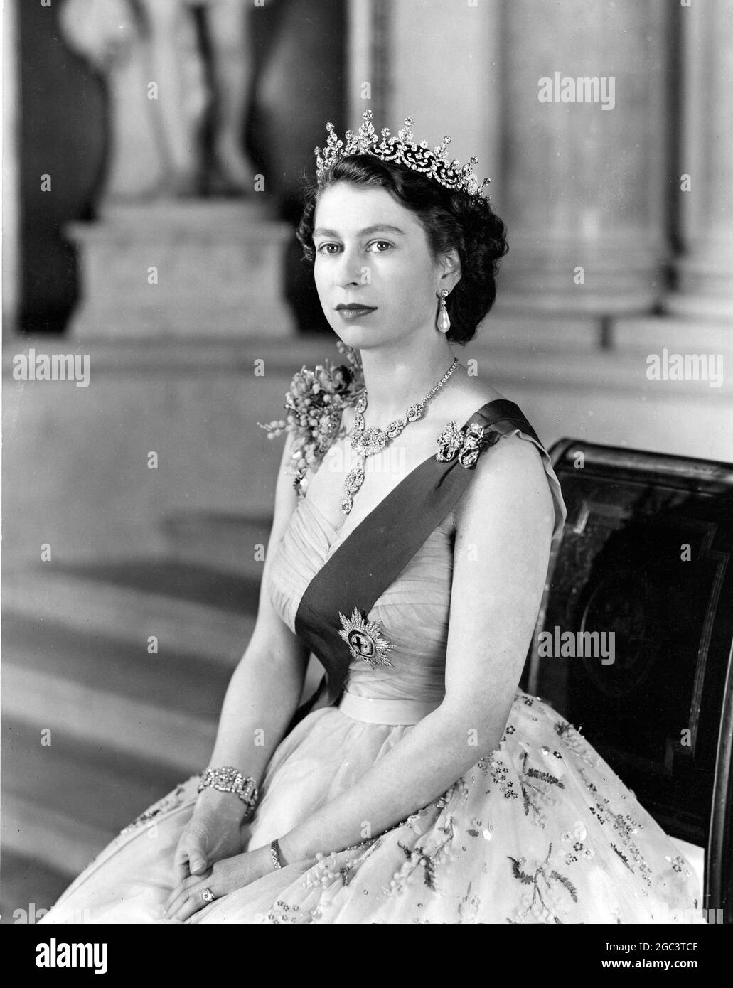 La Reine est vue dans une robe de soirée de tulle jaune ornée de jets de mimosa et de la broderie de paaillette d'or; elle porte le ruban bleu et l'étoile de la jarretière. Voici quelques détails des diamants de sa Majesté présentés dans le portrait: Le collier est un cadeau de mariage de Nizam de Hyderabad; le tiara, également un cadeau de mariage, de la reine Marie. La broche d'arc, les pendants d'oreilles, le bracelet et la montre de poignet en platine sont également en diamants. L'étude de portrait a été prise sur le petit escalier de la Grande entrée à Buckingham Palace. 26 janvier 1954 Banque D'Images
