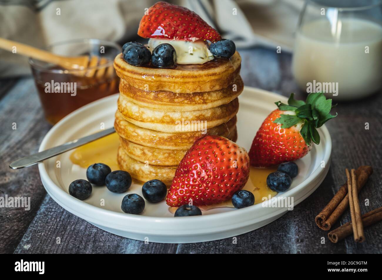 Vue rapprochée de la pile de crêpes avec bâton de miel, fraise, baie bleue et crème fouettée sur l'assiette, délicieux dessert avec lait pour le petit déjeuner. Banque D'Images