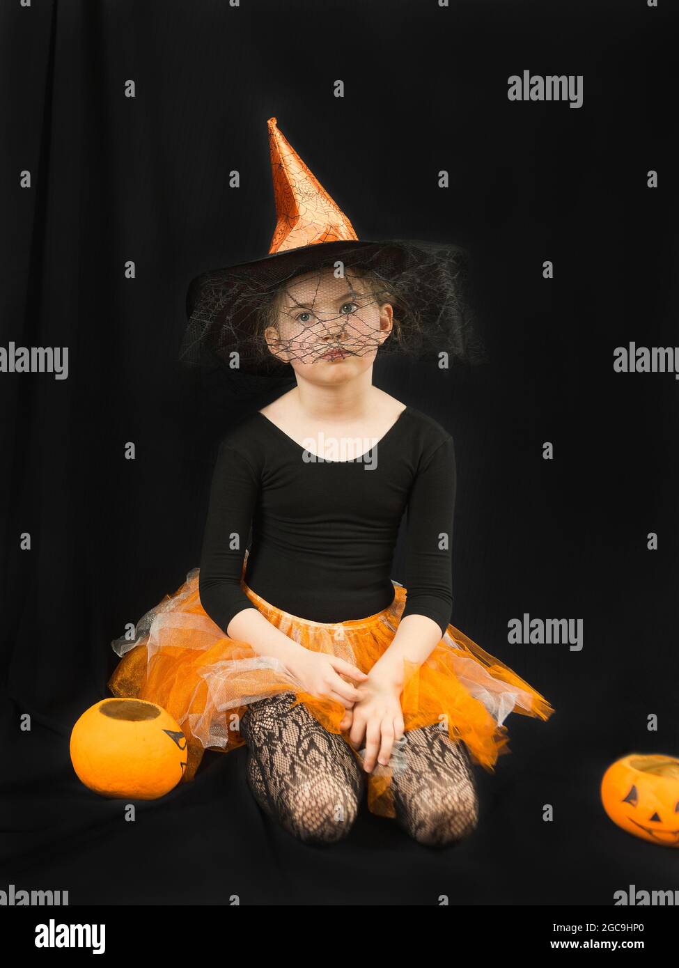 Une fille dans un costume de sorcière - un chapeau avec une toile d'araignée et une jupe orange - se repose avec lassitude sur un fond noir. À côté de lui est un seau-citrouille pour la collecte Banque D'Images