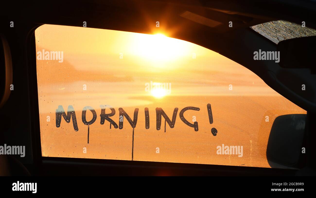 Le mot matin écrit à la main dans la condensation à l'extérieur d'une fenêtre de voiture avec un beau lever de soleil doré sur la côte vue par la fenêtre. Banque D'Images