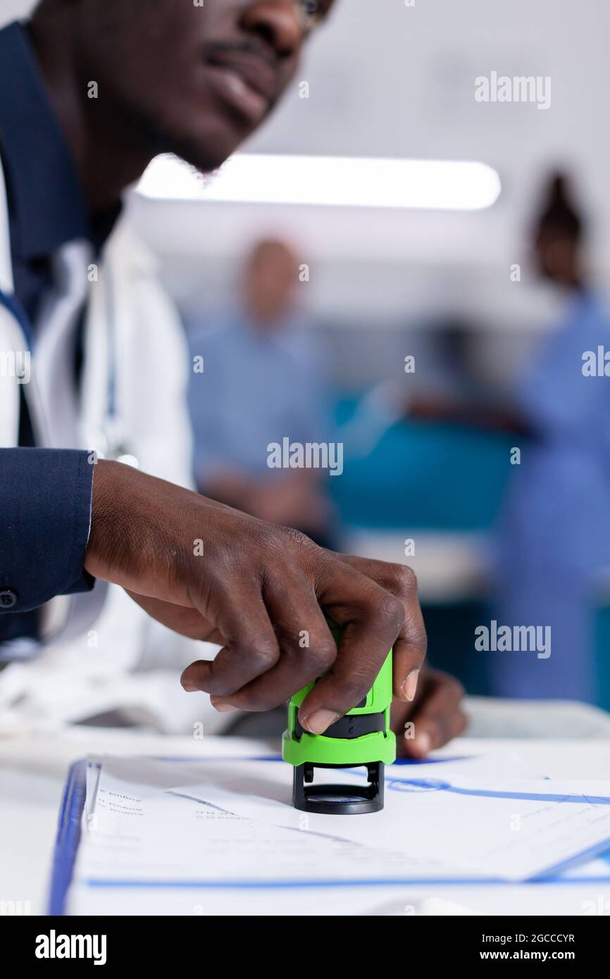Gros plan de la main afro-américaine tenant le timbre sur les papiers pour le rendez-vous de contrôle et la médecine de prescription. Médecin noir utilisant un sceau de bureau professionnel placé dans l'armoire de soins de santé Banque D'Images