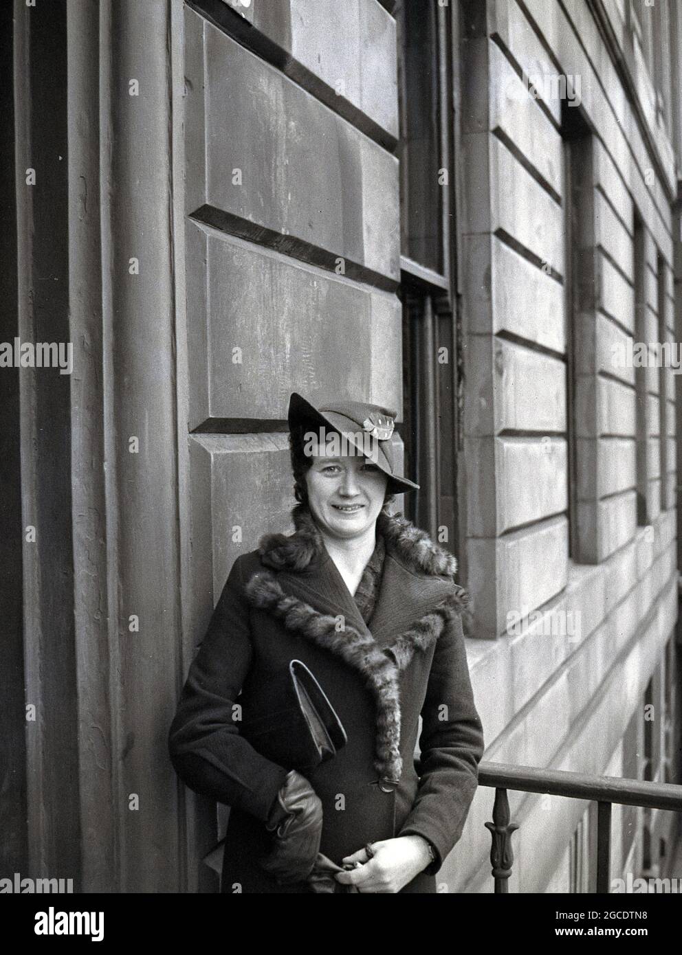 Années 1930, femme historique et élégante portant un chapeau avec une plume  et vêtu d'un manteau, avec des glands décoratifs en fourrure, debout près  d'une entrée sur une terrasse géorgienne, Écosse, Royaume-Uni