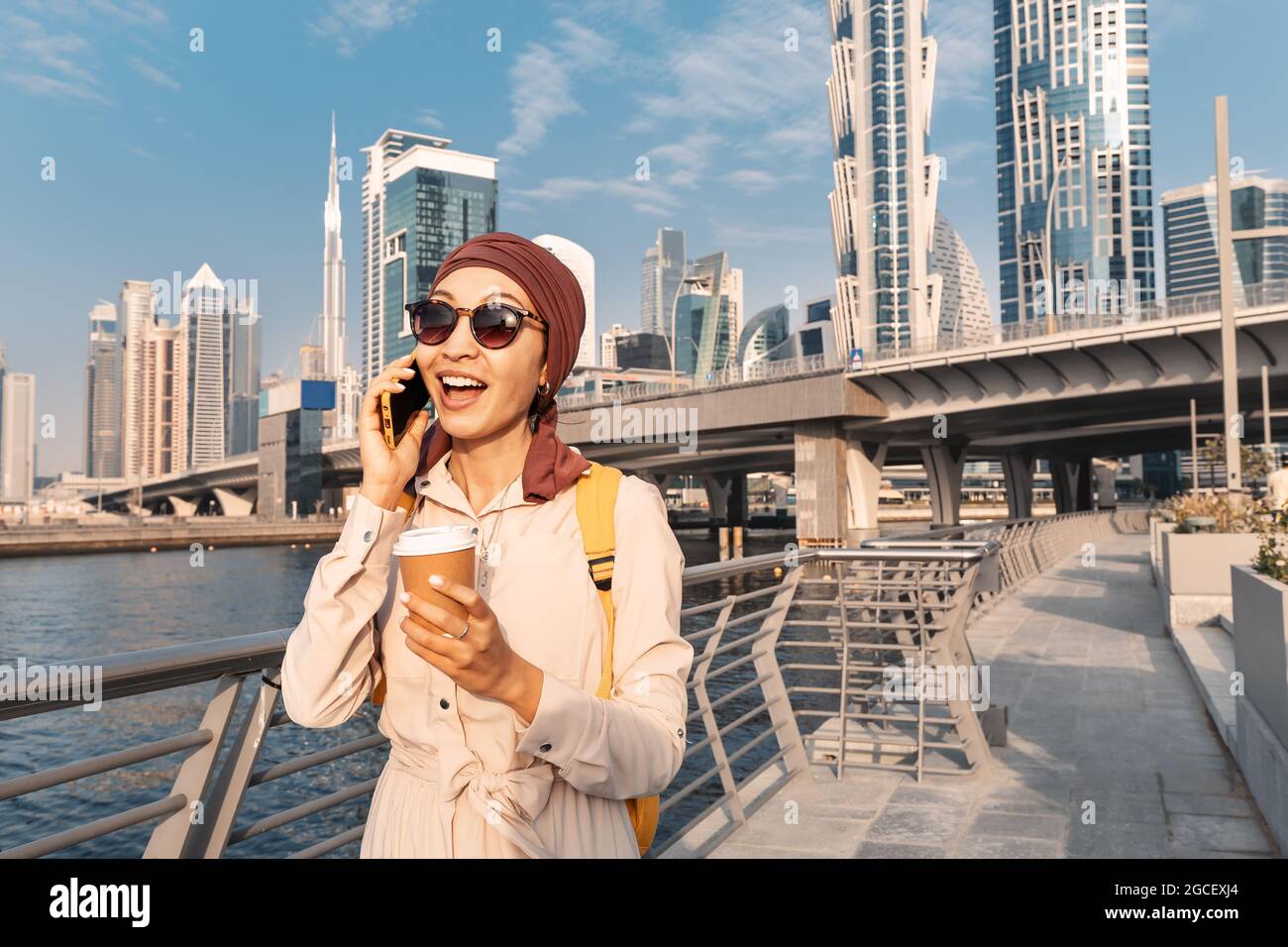 Femme asiatique dans un foulard qui boit du café et parle et commet des commérages au téléphone avec un ami sur le Canal de la promenade de Dubaï Banque D'Images