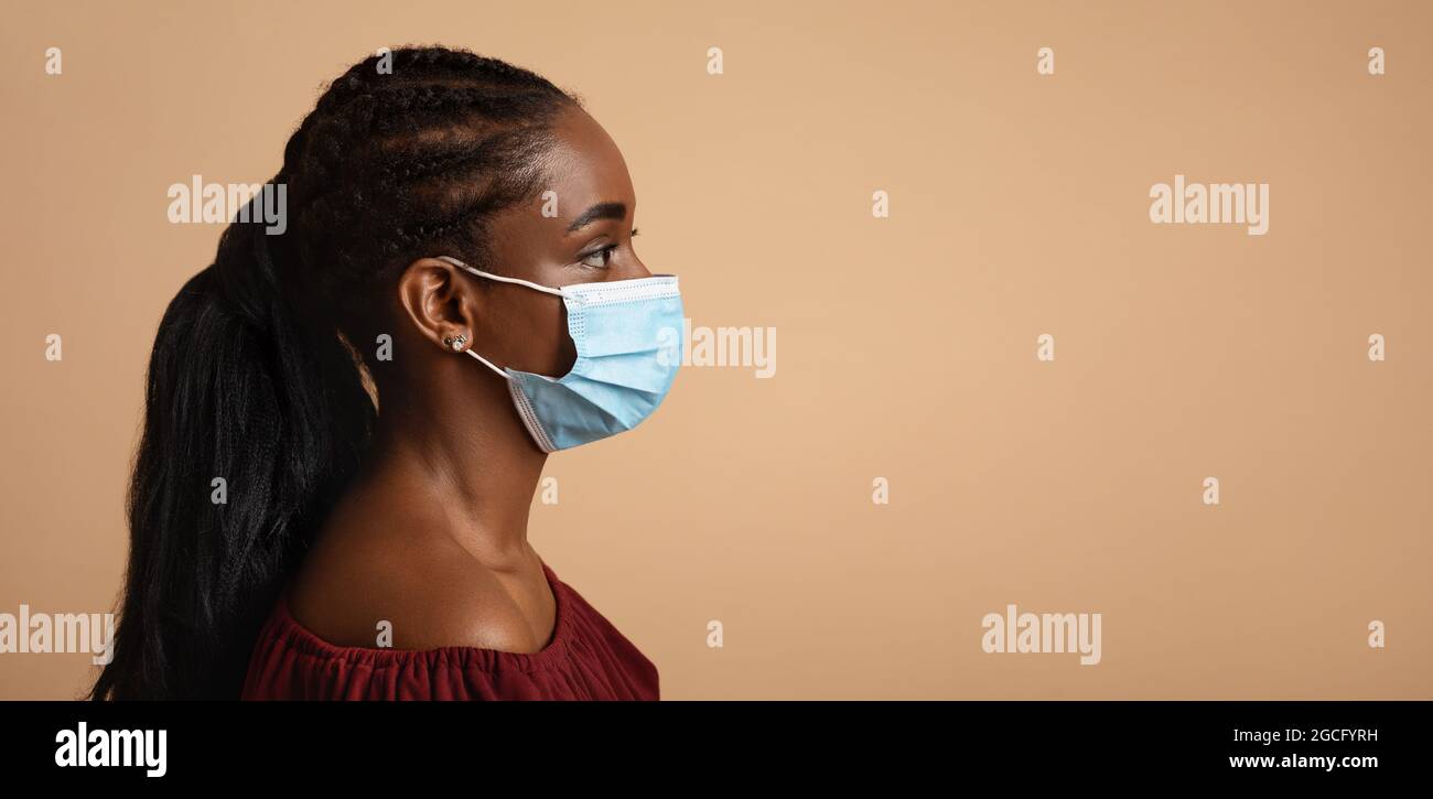 Vue latérale d'une jeune femme noire dans un masque de protection regardant l'espace de copie pour le texte ou la publicité sur fond beige studio, portrait de gros plan, Banque D'Images