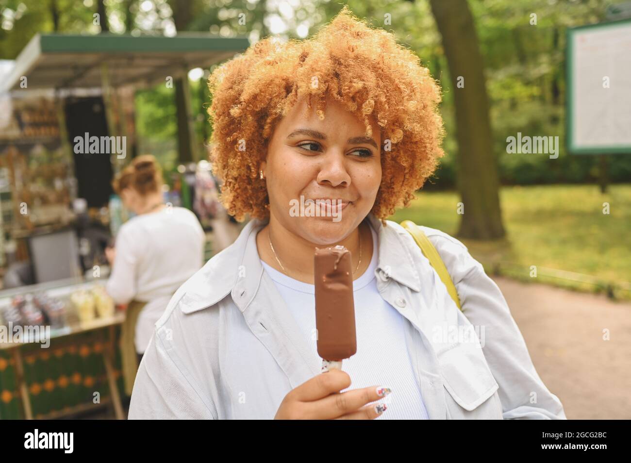 Portrait de style de vie de jeunes souriants naturellement belle et de taille afro-américaine femme avec des cheveux rouges afro manger glace crème chocolat popsicle dans le parc public. Diversité et concept positif du corps Banque D'Images