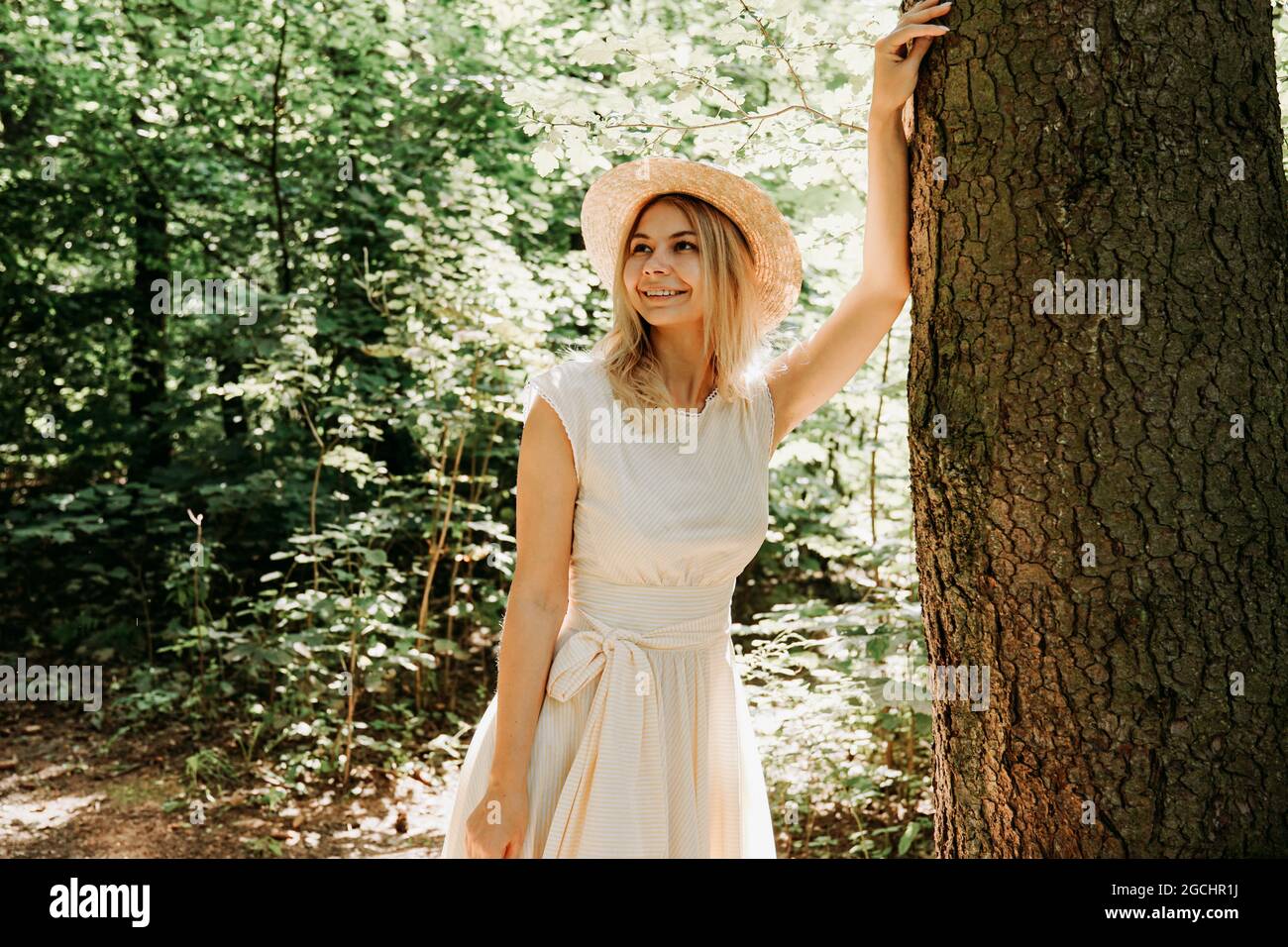 Belle fille dans un chapeau de paille et des supports de vêtements élégants  dans un parc près d'un arbre. Une femme vêtue d'une robe blanche près d'un  arbre. Portrait rapproché de la