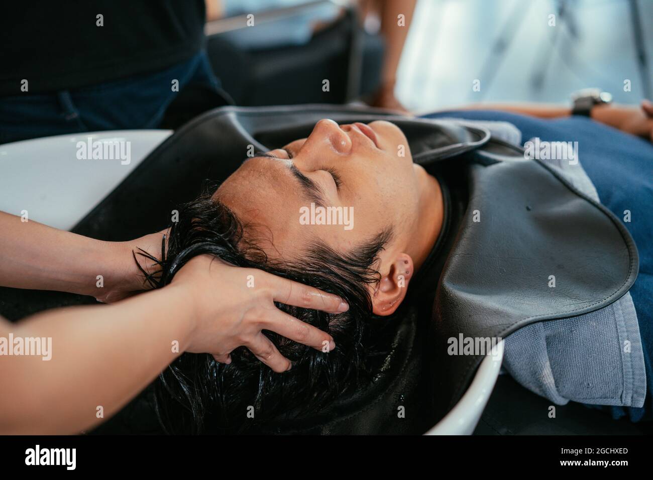 Beau jeune homme asiatique se détendant avec les yeux fermés avec la tête dans l'évier tandis que les mains de la femme de coiffure rinçant les cheveux et les points de pression pressants Banque D'Images