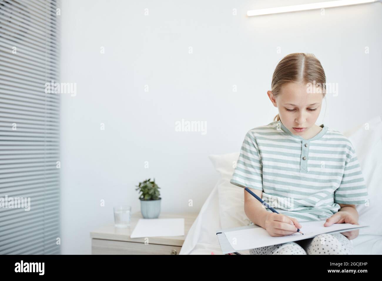 Portrait minimal d'une petite fille assise sur le lit et dessinant dans la salle blanche de l'hôpital, espace de copie Banque D'Images