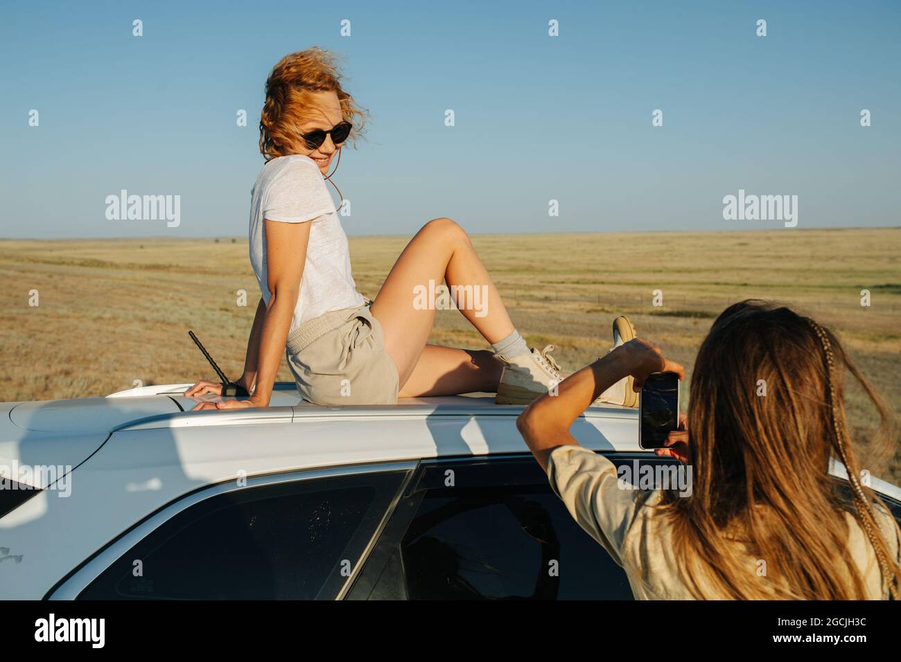 Une femme espiègle assise sur le toit d'une voiture à la campagne, posant pour une photo de sa petite amie en prenant. Belle steppe herbacée en arrière-plan. Banque D'Images