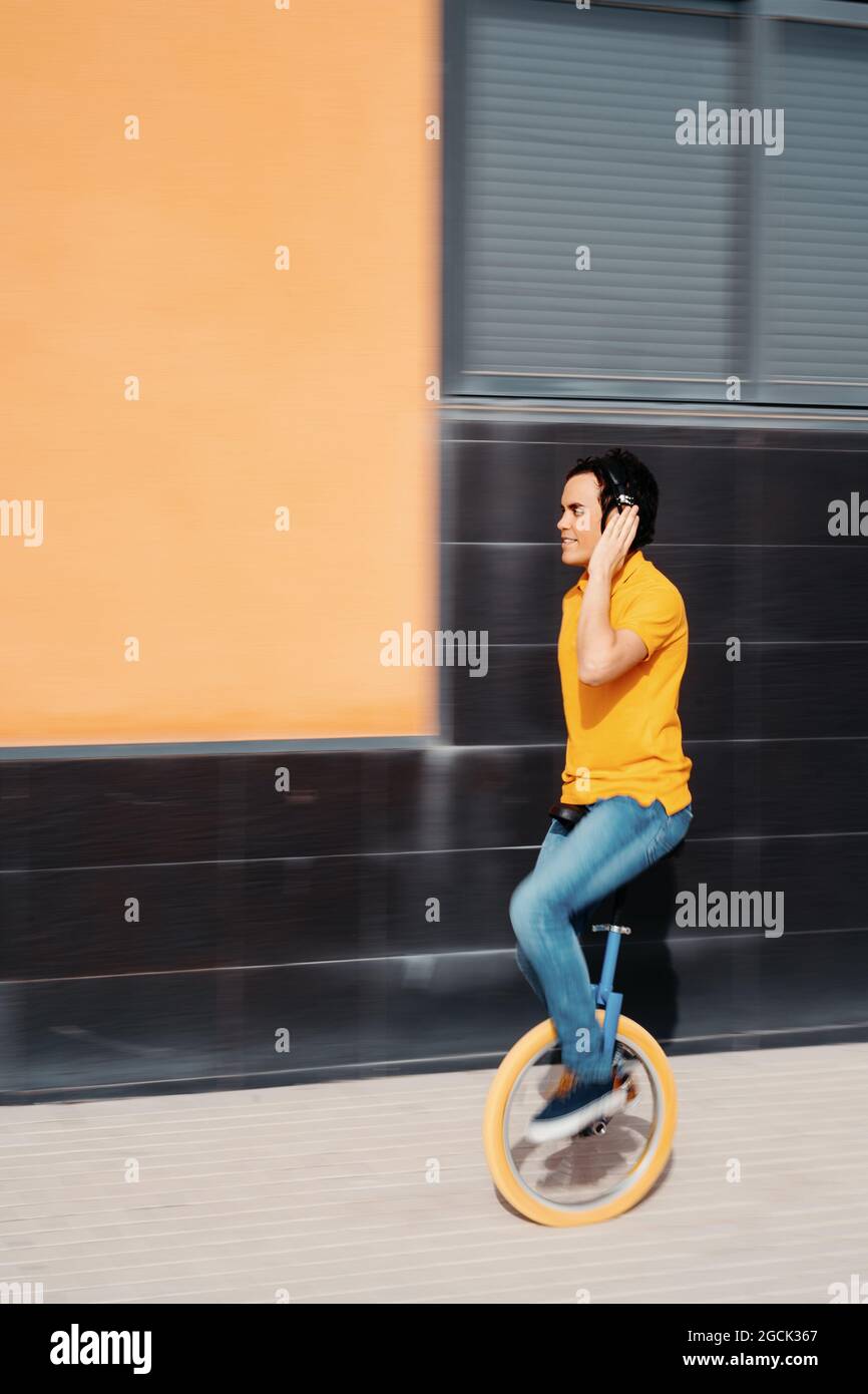 Vue latérale du corps du jeune homme moderne en chemise orange vif et jeans écoutant de la musique à l'aide d'un casque sans fil tout en faisant du monocycle sur le pave Banque D'Images