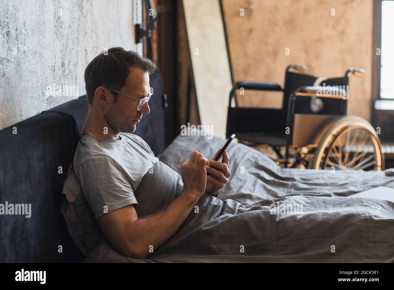 Portrait d'un homme moderne handicapé avec vue latérale, avec un smartphone au lit, un fauteuil roulant en arrière-plan, un espace de copie Banque D'Images