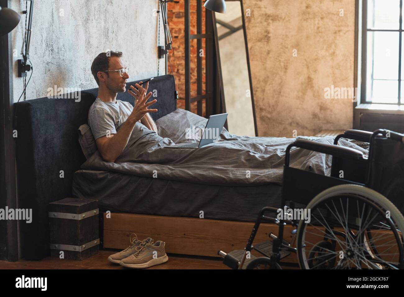 Portrait en vue latérale d'un homme moderne handicapé parlant avec un smartphone dans le lit, fauteuil roulant au premier plan, espace de copie Banque D'Images