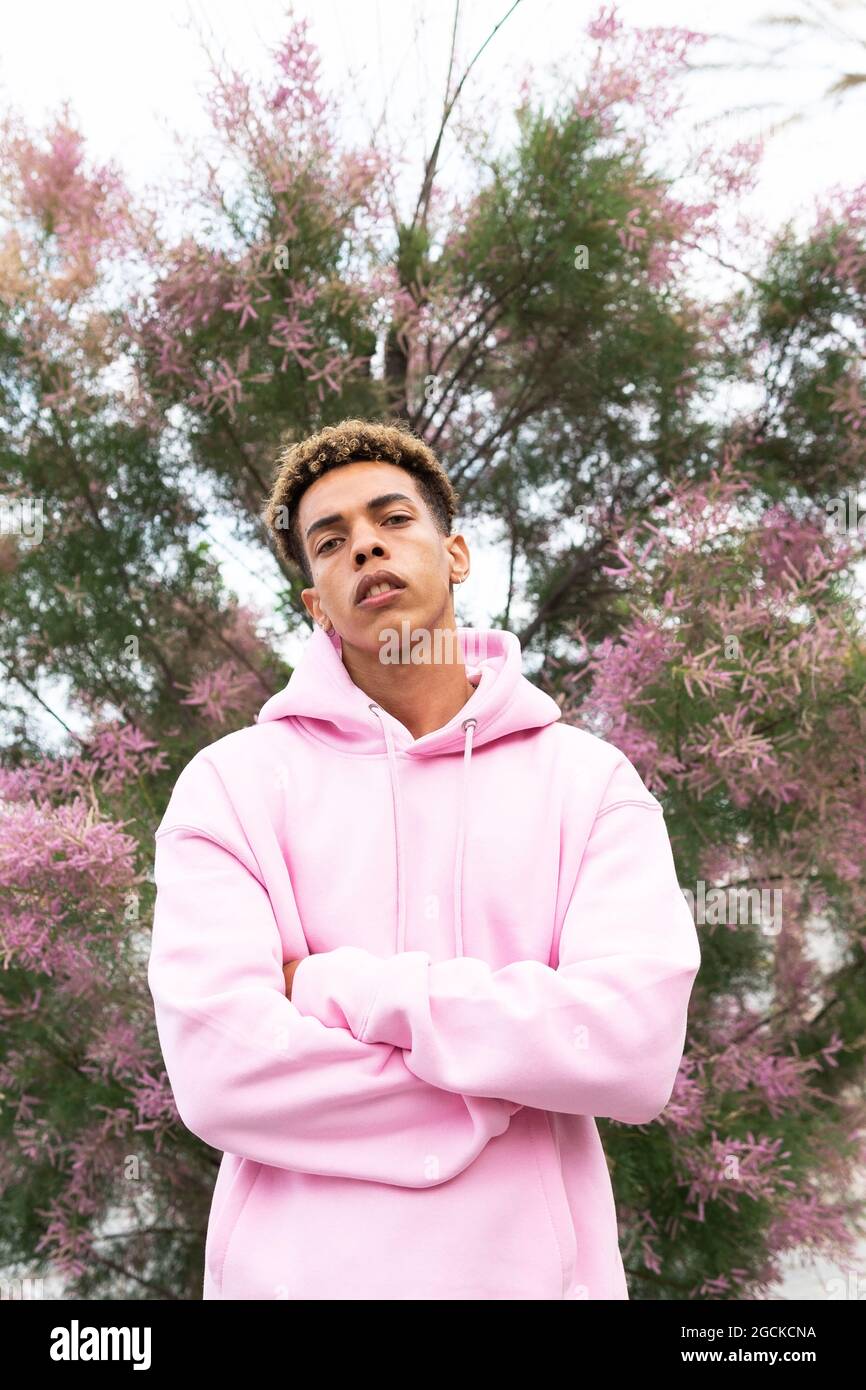 Petit angle de jeune homme ethnique confiant avec une coiffure afro teint  vêtu d'un pull à capuche rose tendance debout avec les bras croisés contre  l'arbre en fleur et le lo Photo