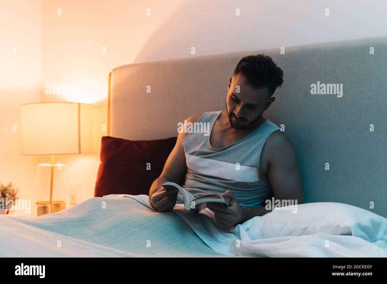 Homme attentionné assis sur un lit sous une couverture et lisant une histoire intéressante dans un livre avant de dormir Banque D'Images