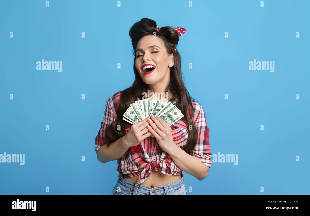 Surjoyeuse jeune femme pinup tenant une grosse somme d'argent près de coeur, excitée par la loterie gagnante sur fond bleu studio. Femme millénaire rétro Banque D'Images