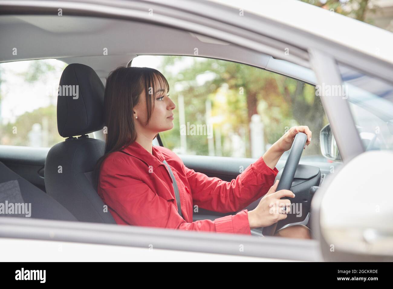 Par la fenêtre vue latérale de la jeune femme confiante en veste rouge conduisant une automobile moderne en ville Banque D'Images