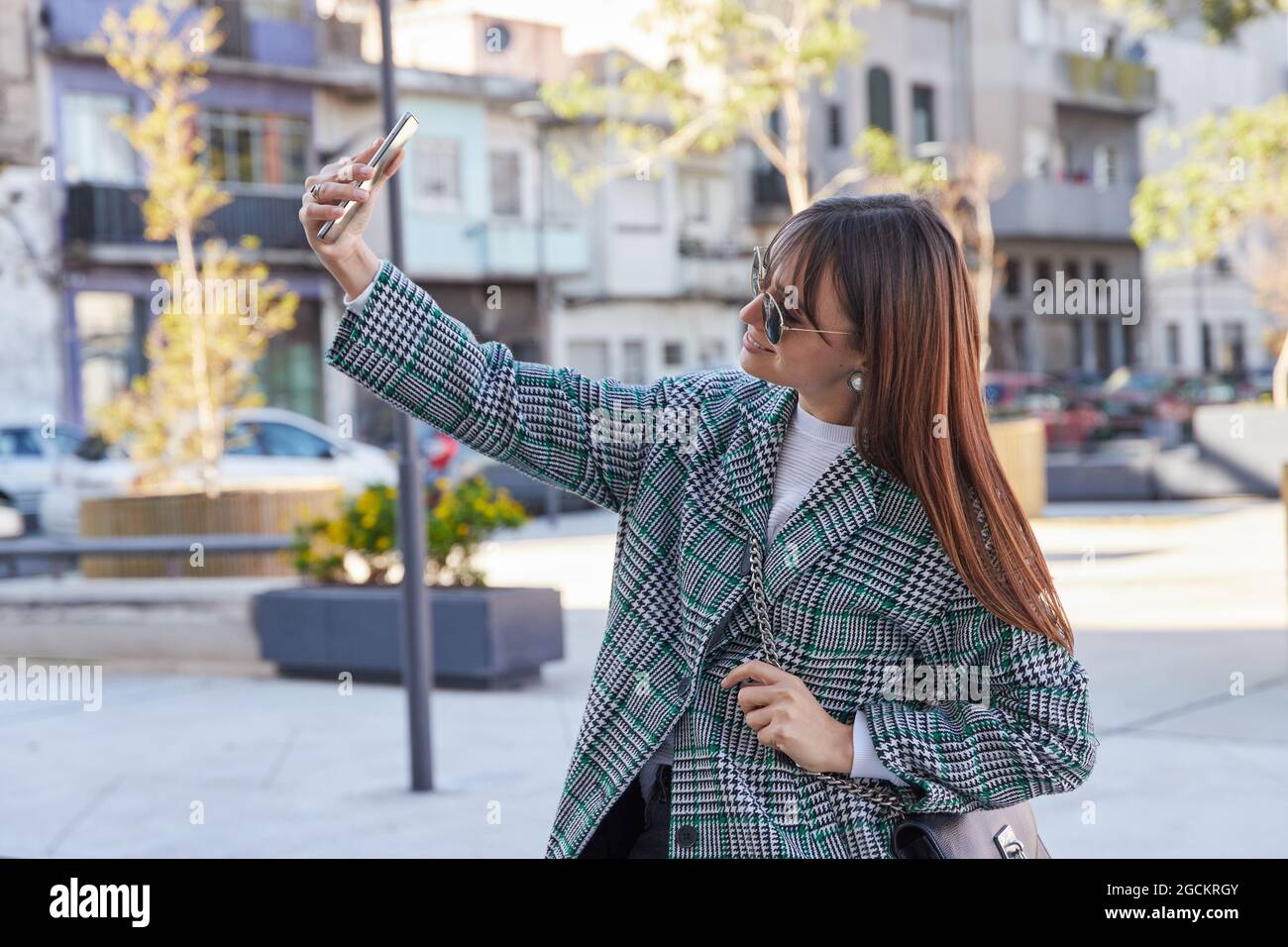 Jeune femme élégante en manteau de pied-de-poule tendance et lunettes de soleil emportant le selfie sur un smartphone tout en se tenant dans la rue de la ville Banque D'Images