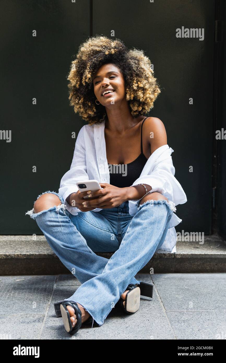 Corps complet de la femme afro americaine positive en jeans dechires et chemise blanche assis avec des jambes croisees et de navigation de telephone cellulaire Photo Stock Alamy