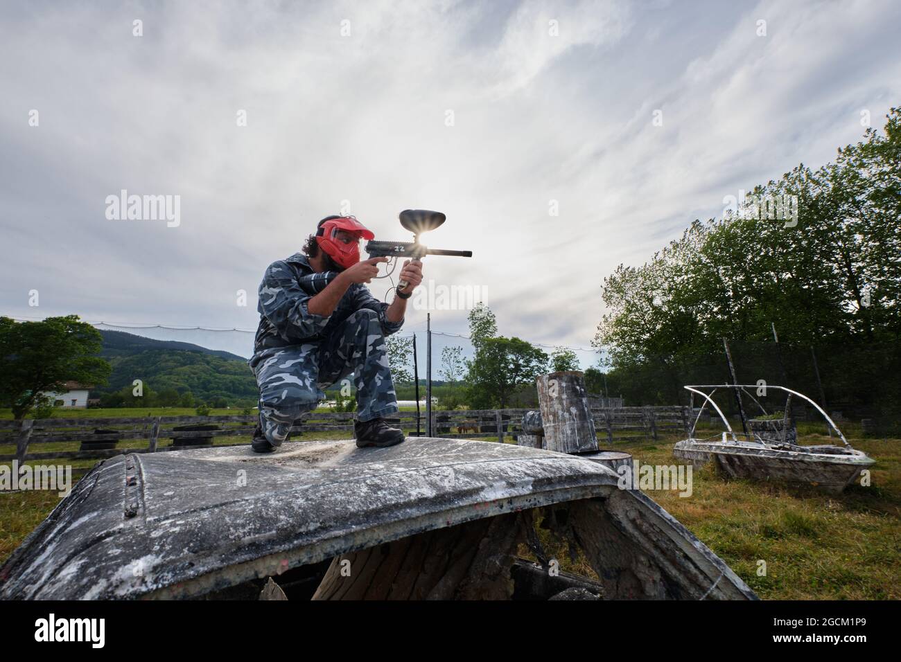 Homme en tenue de camouflage et avec un fusil assis sur une voiture abandonnée et visant tout en jouant au paintball dans la campagne Banque D'Images