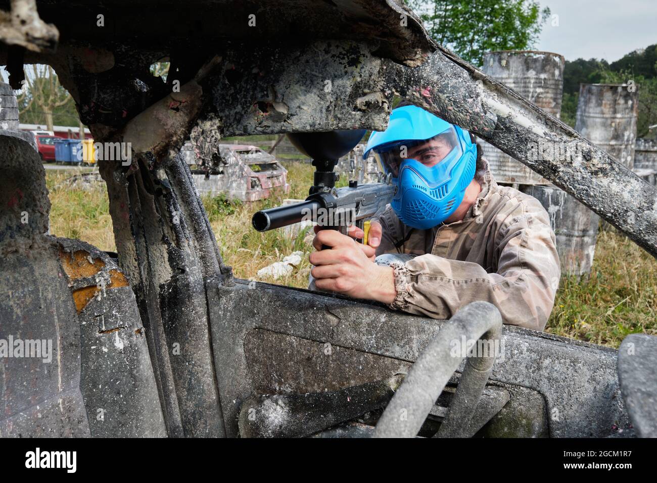 Un joueur masculin dans un casque et avec un fusil jouant du paintball et se cachant derrière une voiture abandonnée de sorbby pendant le match Banque D'Images