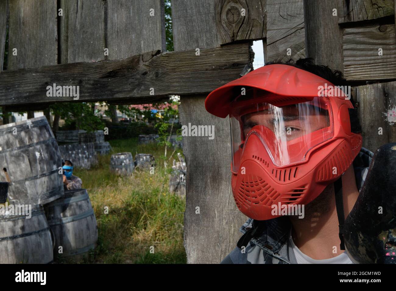 Un joueur masculin dans un masque et avec un pistolet à paintball debout derrière une clôture en bois pendant le match Banque D'Images