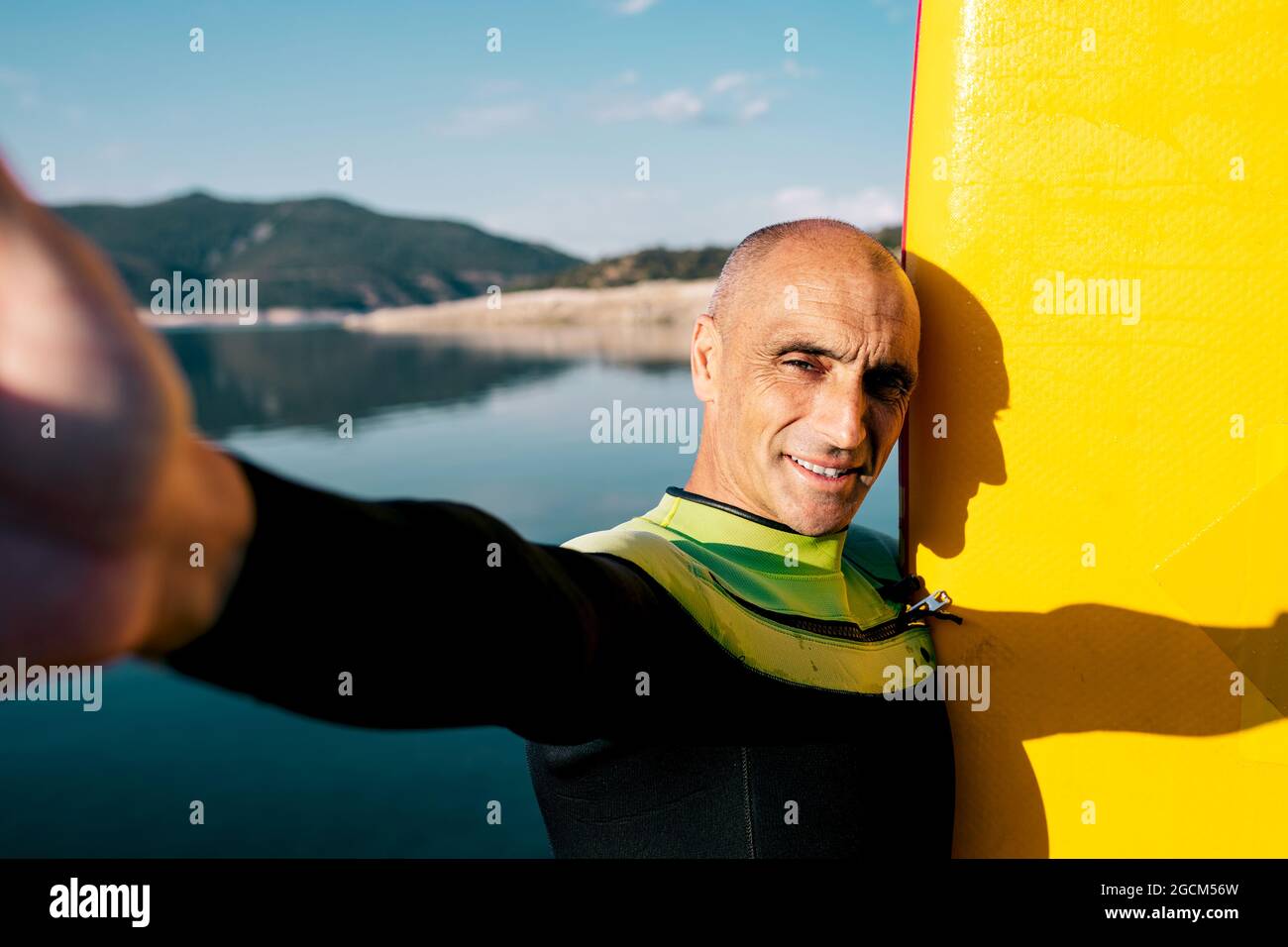 Homme actif positif d'âge moyen en combinaison debout près du paddle board et regardant l'appareil photo tout en prenant autoportrait contre le lac calme en été da Banque D'Images