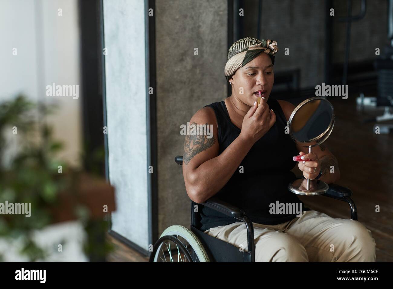 Portrait d'une femme afro-américaine en fauteuil roulant mettant sur un rouge à lèvres tout en tenant le miroir à l'intérieur de la maison Banque D'Images